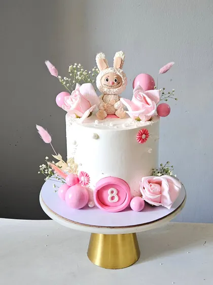 A chic and modern cake featuring a Labubu character topper. The design is finished with delicate pink roses, dried grasses, and decorative spheres for a trendy, "pinteresty" look.