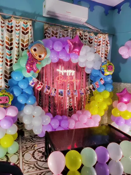 A Cocomelon themed birthday decoration with a pink shimmer curtain and a pastel balloon arch. This setup includes a photo banner with pictures of the birthday child.