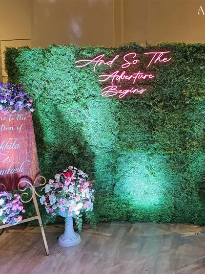 A creative photo backdrop featuring a lush green moss wall with a neon sign that reads "And So The Adventure Begins". This modern and fun detail is perfect for capturing memories.