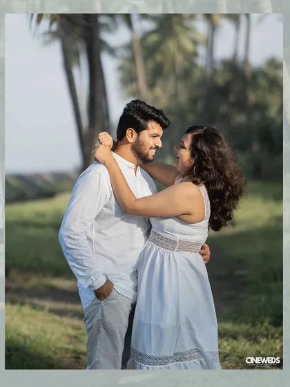 A sweet, candid pre-wedding photo of a couple in white, sharing a look of love amidst a grove of palm trees.