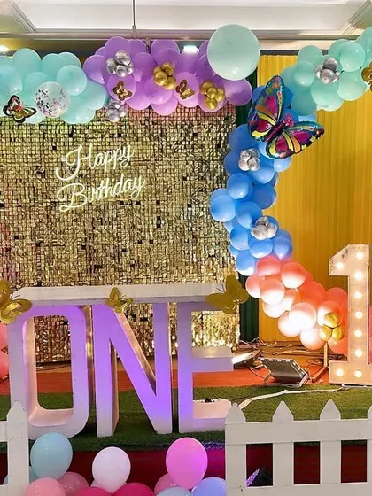 A pastel-colored butterfly theme for a first birthday. This decor features a gold shimmer wall, a "ONE" table, and a beautiful rainbow-colored balloon arch.
