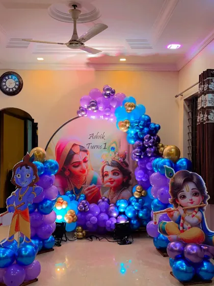 A beautiful Krishna-themed first birthday decoration for a home setting. The setup includes a custom backdrop, character cutouts, and a vibrant blue and purple balloon arch.