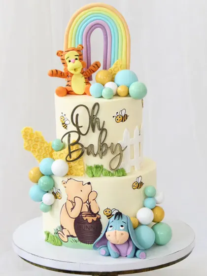 A sweet and classic Winnie the Pooh themed cake, perfect for a baby shower. This two-tiered design features Pooh, Tigger, and Eeyore, along with a pastel rainbow and buzzing bees.