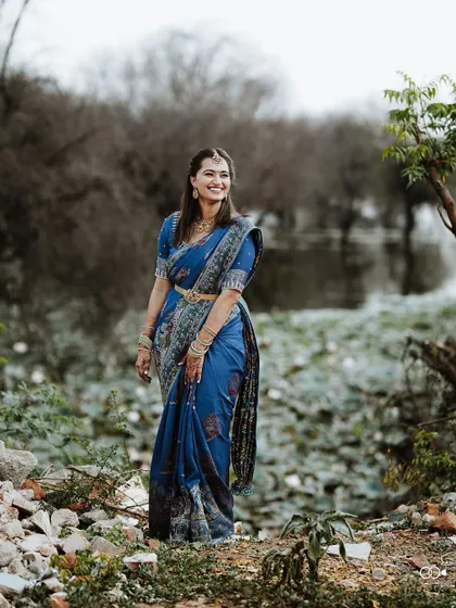 A beautiful portrait of the bride in a vibrant blue saree, standing gracefully in a natural, rustic setting.