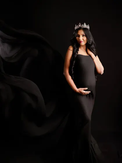 A dramatic and regal portrait. The mom-to-be wears a crown and a flowing black gown, creating a powerful and elegant image against a dark background.