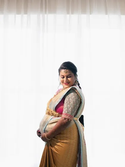 A classic maternity portrait of a woman in a golden saree, framed by lamps and soft window light, showcasing her radiant pregnancy glow.