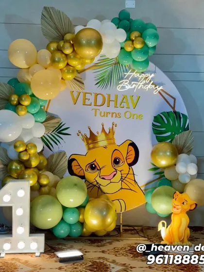 A vibrant Lion King "Wild One" birthday setup featuring a crowned Simba on a circular backdrop. The decor is accented with a "Happy Birthday" neon sign, a large light-up number '1', and an organic balloon garland in gold and green tones.