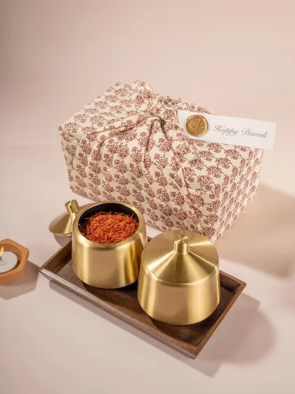 A minimalist festive gifting scene for Doro, featuring brass containers and a fabric-wrapped gift. The soft, neutral color palette creates a calm and sophisticated holiday mood.