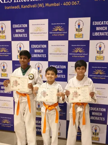 Three proud students with their new orange belts and certificates after a successful grading at KES International School.