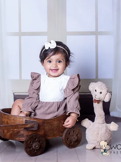 A sweet baby girl with a big smile, sitting in a wooden wagon next to a stuffed animal. The simple, clean background with a window light effect creates a classic and timeless portrait.