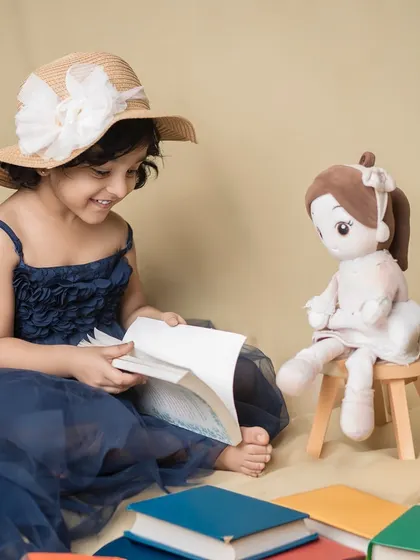 Story time with a special friend. A little girl in a blue dress and hat reads a book to her doll, surrounded by a sea of colorful books.