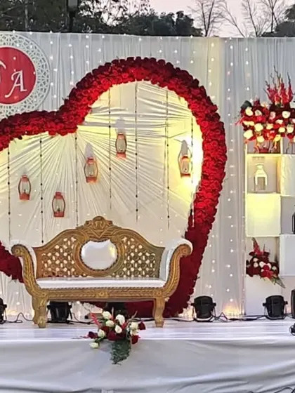 A romantic engagement stage design featuring a large heart-shaped floral arch made of red roses. The couple's initials are displayed above, and the white paneling with shelving for decor adds a personalized touch.
