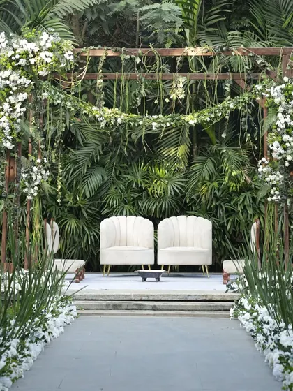 A simple yet elegant ceremony stage with a wooden frame adorned with white flowers and greenery, set against a natural backdrop of lush foliage.
