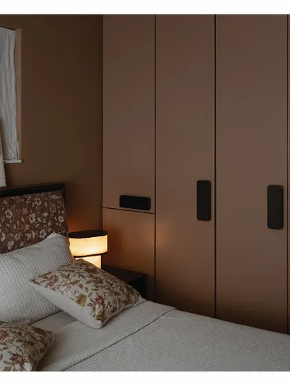 The master bedroom in our 'Home' project is a desert-toned, cozy space. We designed these full-length wardrobes in a mauve finish with bespoke emerald-toned handles, complementing the maroon headboard and taupe walls.