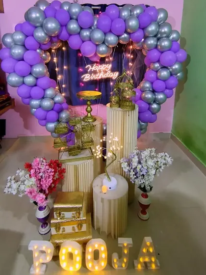 A midnight surprise birthday decoration with a heart-shaped balloon arch in purple and silver, complete with fairy lights and flowers.