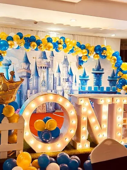 A royal prince-themed first birthday party. The stage features a large castle backdrop, "ONE" marquee letters, and a blue and yellow color scheme.