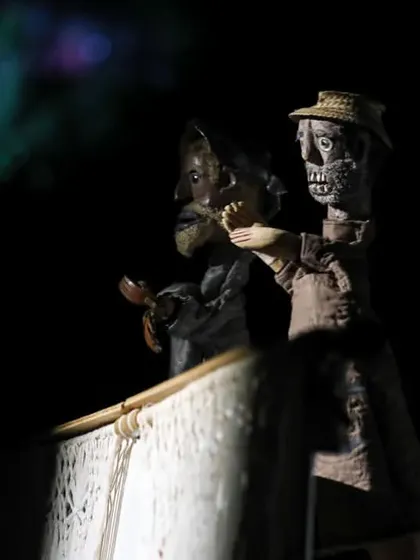 Two skeletal-looking puppets interact in a darkly lit scene from 'Mamulengo de la Mancha', showcasing the show's witty and satirical tone.