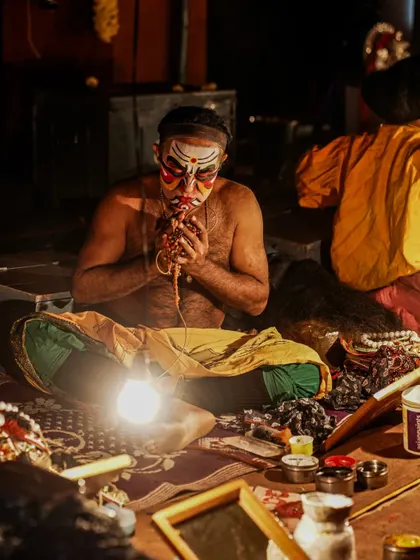 A close-up of a Yakshagana artist applying his 'bannagarike' or traditional makeup. The light from a single bulb illuminates his focused expression. These quiet, transformative moments backstage are just as important to the story as the performance itself.