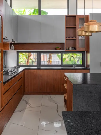 The kitchen in the SAMAM House is designed for functionality and connection to the outdoors. A wide window over the sink provides natural light and garden views, while the material palette of teak, grey cabinetry, and granite countertops ensures durability.