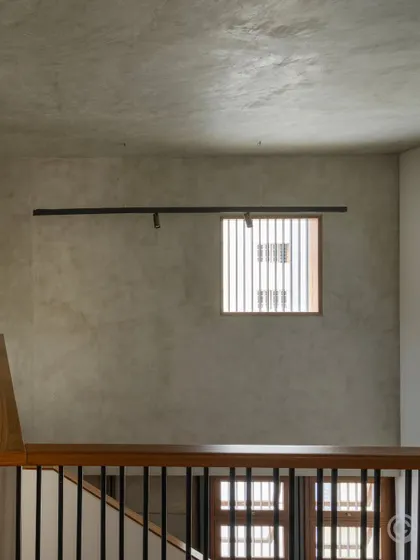 A minimalist composition showing the texture of a hand-finished concrete wall, punctuated by a simple track light and a wood-framed window with vertical slats. This highlights our appreciation for raw, unadorned surfaces.