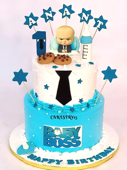 A sharp two-tier Boss Baby cake for a first birthday. The design features a fondant tie, cookies, a milk bottle, and star toppers with the baby's name.