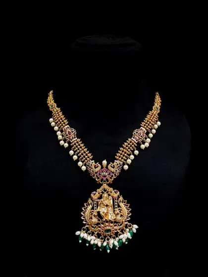 A long temple necklace, or haram, with a detailed Lakshmi pendant and green bead drops. This is a statement piece, perfect for a bride on her wedding day.