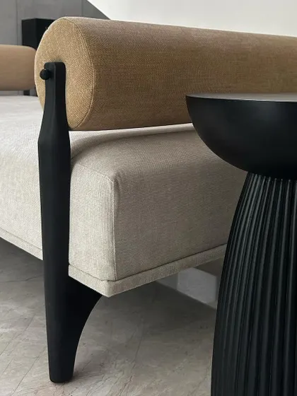 A detail shot of the day bench, showing the contrast between the smooth, dark-stained teak leg and the textured semi-suede upholstery of the main body and bolster pillow.