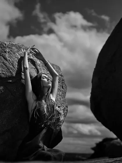An outdoor fine art portrait that tells a story of solitude and strength. Shot in black and white against a dramatic sky and rugged rocks, this image emphasizes form, texture, and the powerful presence of the model in nature.