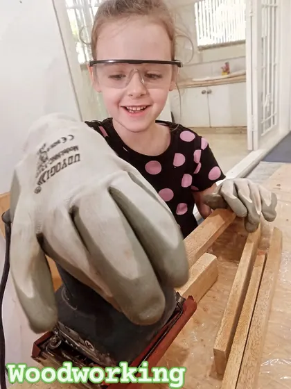 Look at that smile! She's having a great time learning how to use a power sander, an important skill in our woodworking workshops for kids.