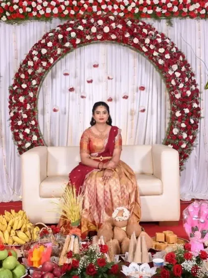 A bride seated at her engagement ceremony, with a circular red rose arch behind her and a traditional offering of fruits and coconuts in the front.