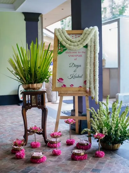 A beautiful welcome setup for a Varamahalakshmi puja. The personalized sign is framed with jasmine garlands, and the entrance is decorated with lotus flowers and traditional lamps, creating a serene and auspicious ambiance.