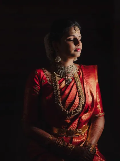 A dramatic bridal portrait with chiaroscuro lighting, highlighting the rich red of her saree and the sparkle of her jewelry against a dark background.