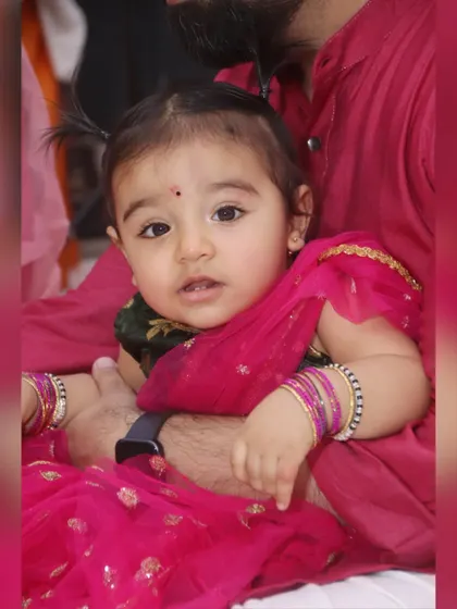 A close-up portrait of a baby girl in a traditional pink outfit, looking inquisitively at the camera.