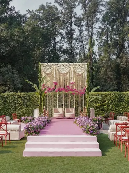 The mandap for a garden wedding, featuring a backdrop of traditional Indian 'jaal' work and a cascade of flowers. The mix of red chairs and pastel decor creates a unique, eclectic look.