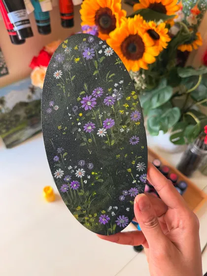 A close-up of the finished oval wildflower painting, held against a background of fresh sunflowers. You can see the delicate purple, white, and yellow flowers I painted.