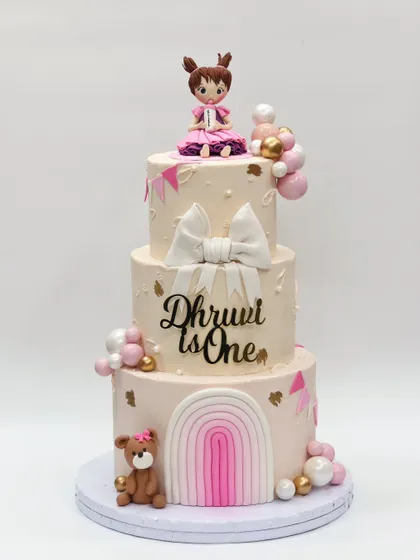 A grand three-tier first birthday cake with a soft pink and white theme, featuring a large bow, a rainbow, a teddy bear, and a girl figurine.