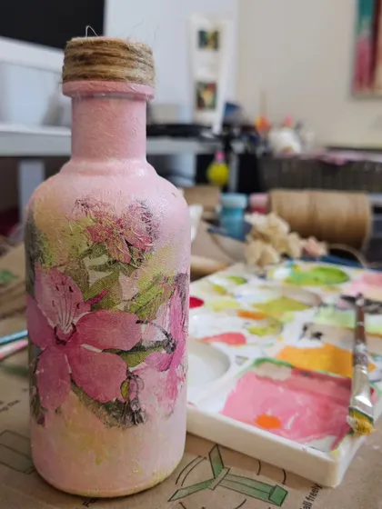A finished decoupage bottle decorated with soft pink textures and floral designs, sitting on my messy-but-loved workspace, demonstrating my custom decor services.