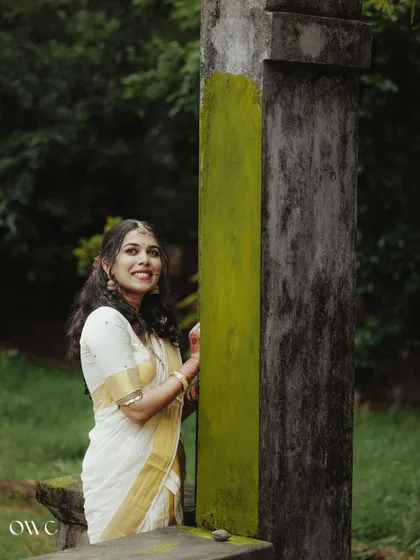A serene portrait of a bride in a kasavu saree, leaning against a moss-covered pillar.
