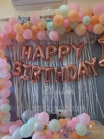 A beautiful pastel-themed decoration for a first birthday at home. The wall is decorated with a white fringe curtain, rose gold "Happy Birthday" letters, and a balloon arch in soft pastel shades.