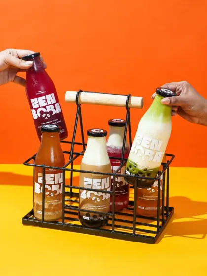 Two hands reach for bottles of Zen Boba from a stylish metal and wood carrier. This lifestyle shot against a bright, two-toned background conveys a sense of sharing and modern convenience.