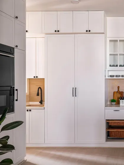 This view highlights the smart storage solutions in this white kitchen, including a tall pantry unit and integrated appliances. The black hardware adds a touch of modern contrast.