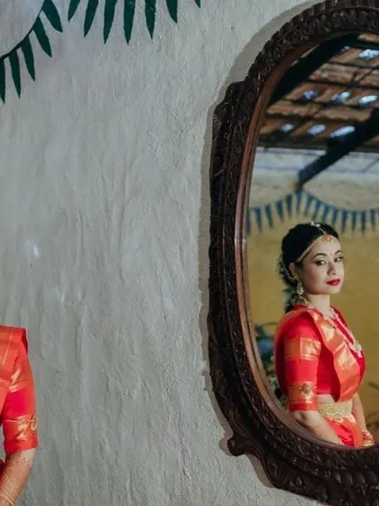 A creative mirror shot reflecting the bride's elegant profile. Her makeup is kept classic with a bold red lip, perfect for her vibrant red saree.