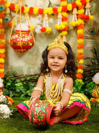 A sweet little Radha with curly hair and a crown, posing with her makhan pot.
