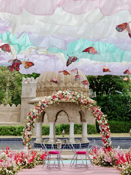 A magical, spring-inspired Mehendi theme. The couple's seating is set before a floral arch, with a dreamy sky of pastel ruffled drapes and fluttering butterflies creating a truly whimsical atmosphere.