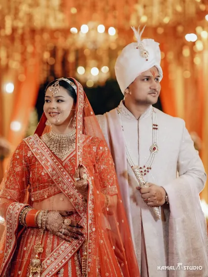 A classic portrait of the couple, Yogesh and Reena, in their wedding attire, standing against a warm, festive backdrop.