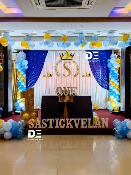 A royal first birthday celebration with a prince theme, using a blue and gold color scheme, crown motifs, and a grand stage with draped curtains.