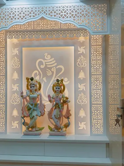 A pristine white Corian mandir featuring intricate jali work on the sides and top arch. The back panel is subtly engraved with religious symbols and a large Ganesha outline, all softly illuminated to create a tranquil and sacred space.