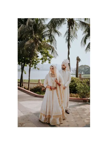A regal, full-length portrait of the couple. Their elegant white and gold outfits stand out against the lush greenery and lakeside background of the luxury venue.