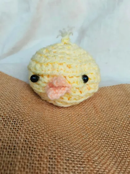A close-up product shot of the chick amigurumi on a textured burlap surface. This shows the details of its little beak and eyes.