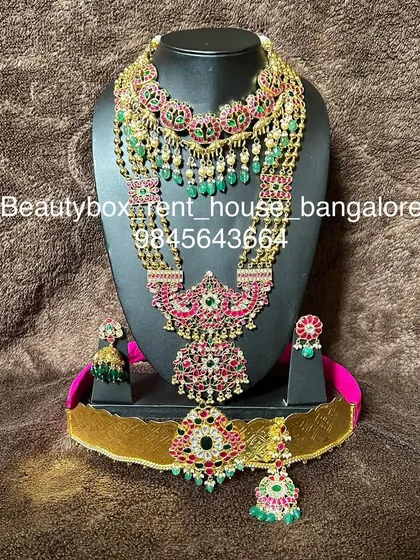 A stunning Jadau-style bridal set with red and green stones and green bead drops. This rental includes multiple necklaces, earrings, maang tikka, and a vaddanam.